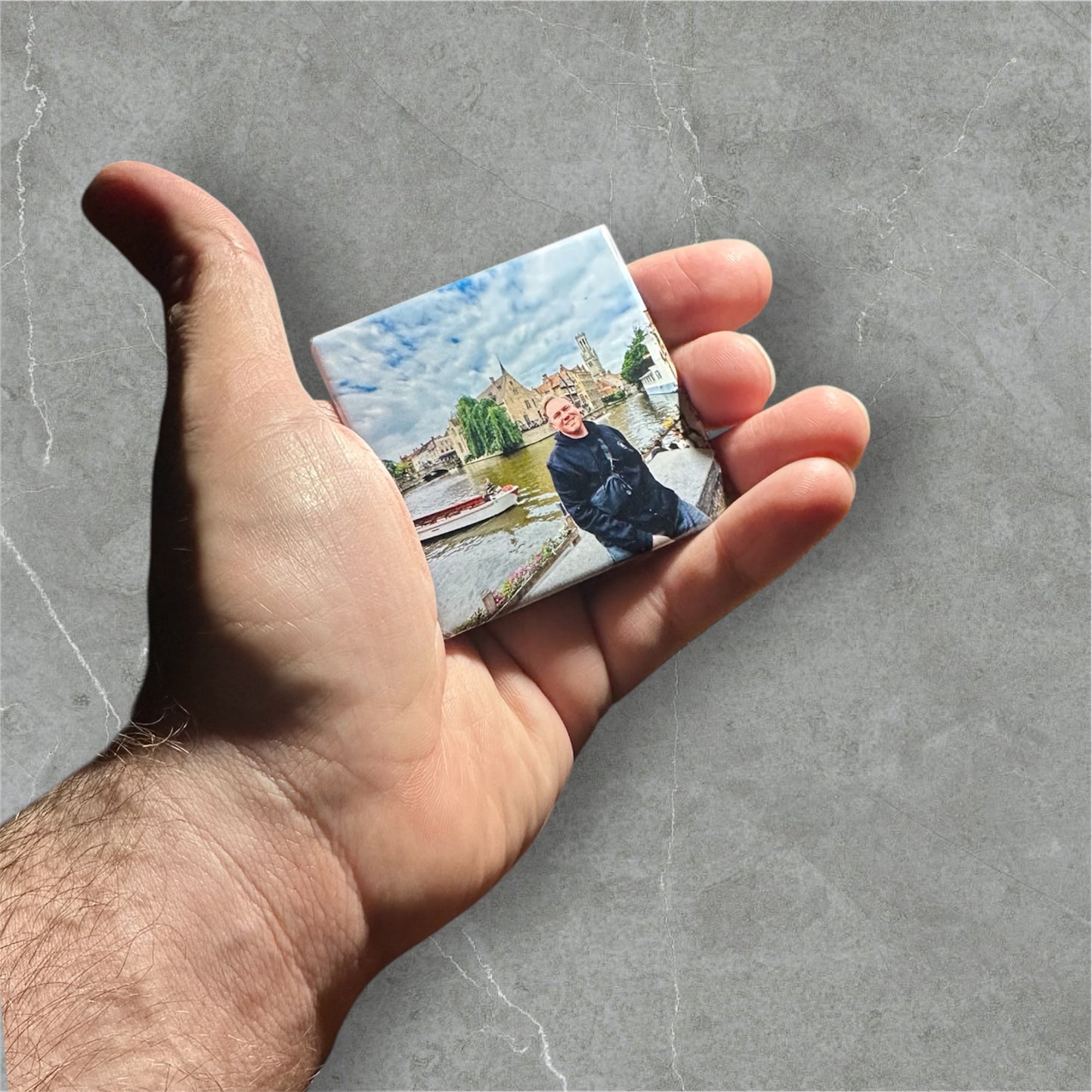 Hand holding a square magnet of a landscape with a person on a gray surface