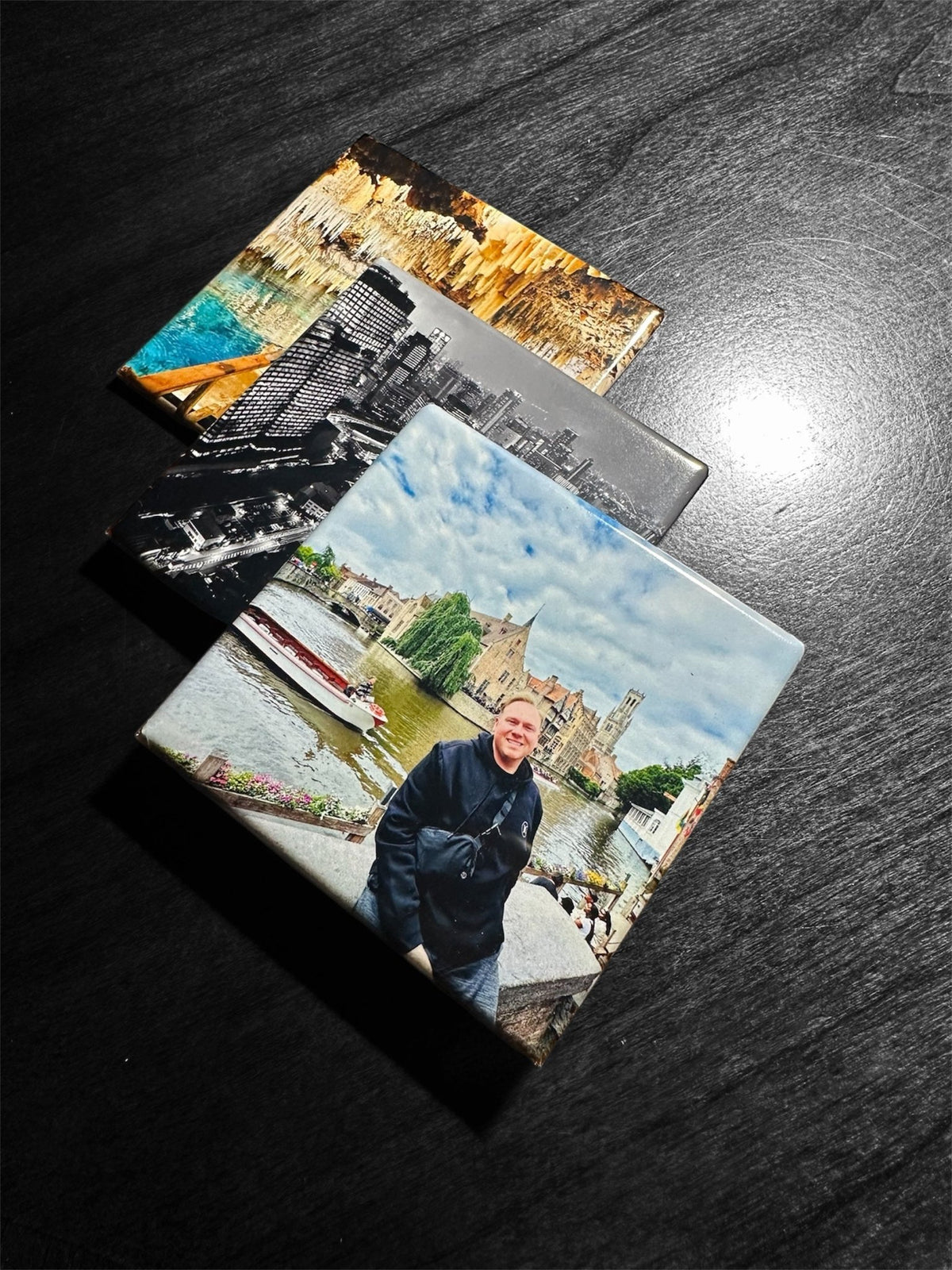 Three square photo magnets with printed scenic and portrait images, arranged diagonally on a dark wood surface.