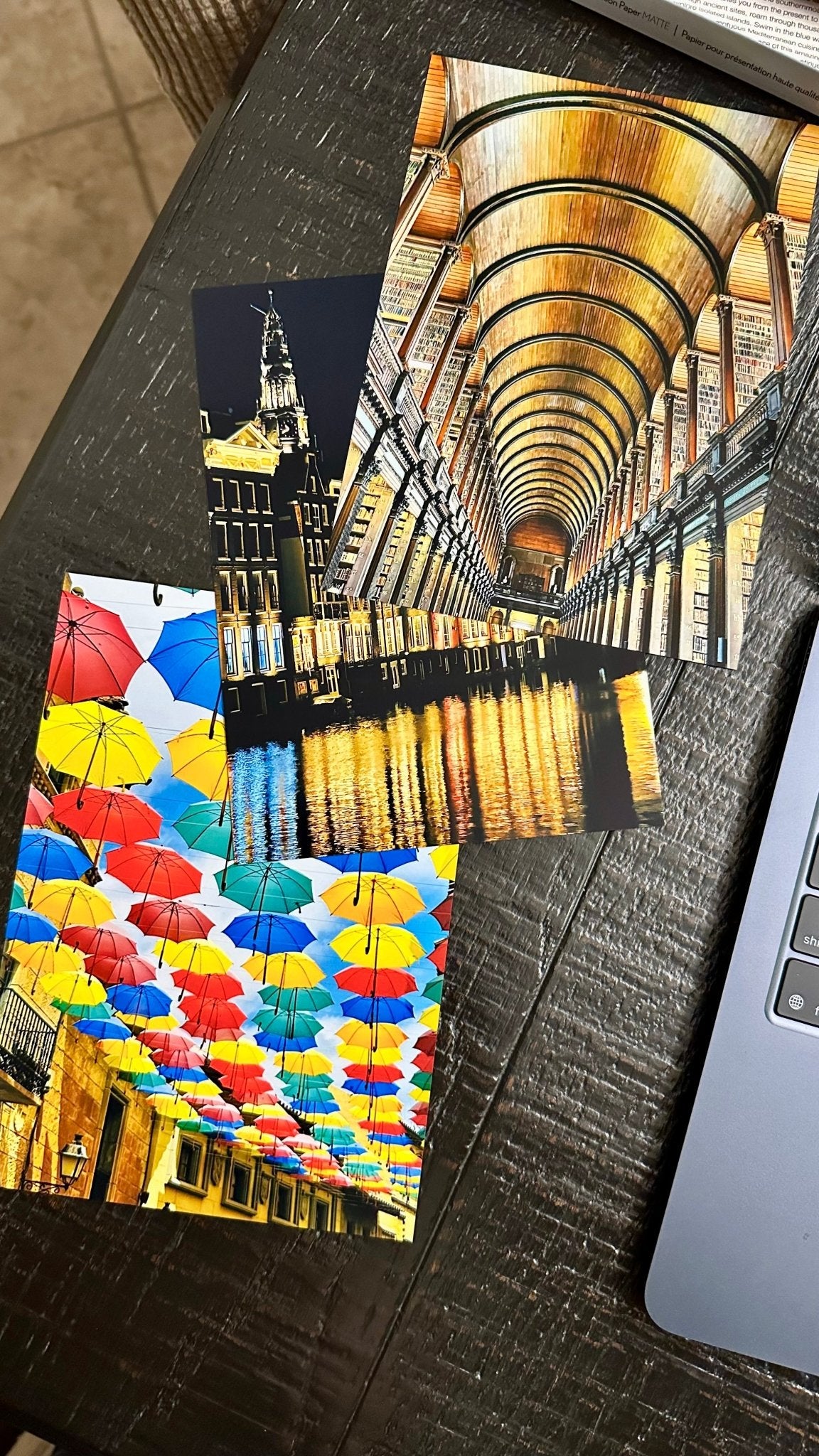 Three printed art cards or postcards are arranged on a dark wooden table next to a laptop. The top card shows a perspective view of a grand arched hall with warm golden reflections.