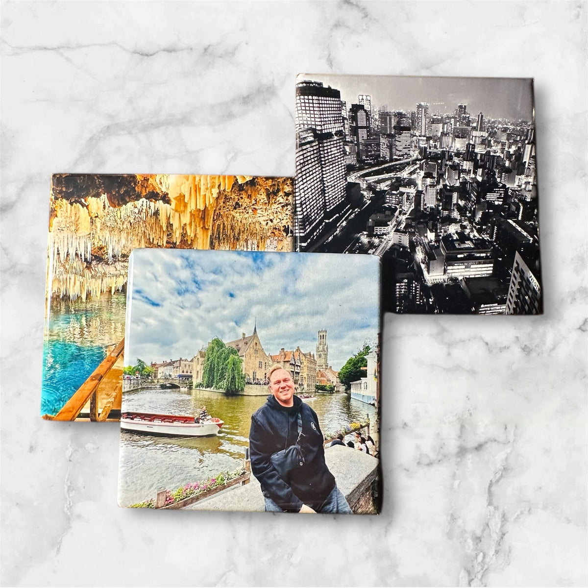 Three square photo magnet prints with scenic and architectural photography displayed on a white marble surface. One print shows a black-and-white city skyline, another depicts a colorful cave formation over water, and the third shows a man standing by a river with historic buildings in the background.